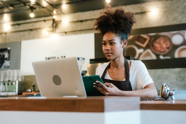 Barista in dark-brown apron working on green tablet and silver laptop at wooden counter inside coffee shop